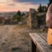 Primo piano delle mani di un musicista che suona una chitarra acustica all'aperto durante il tramonto, con rovine antiche sfocate sullo sfondo, a simboleggiare la connessione tra musica, spiritualità e il momento presente.